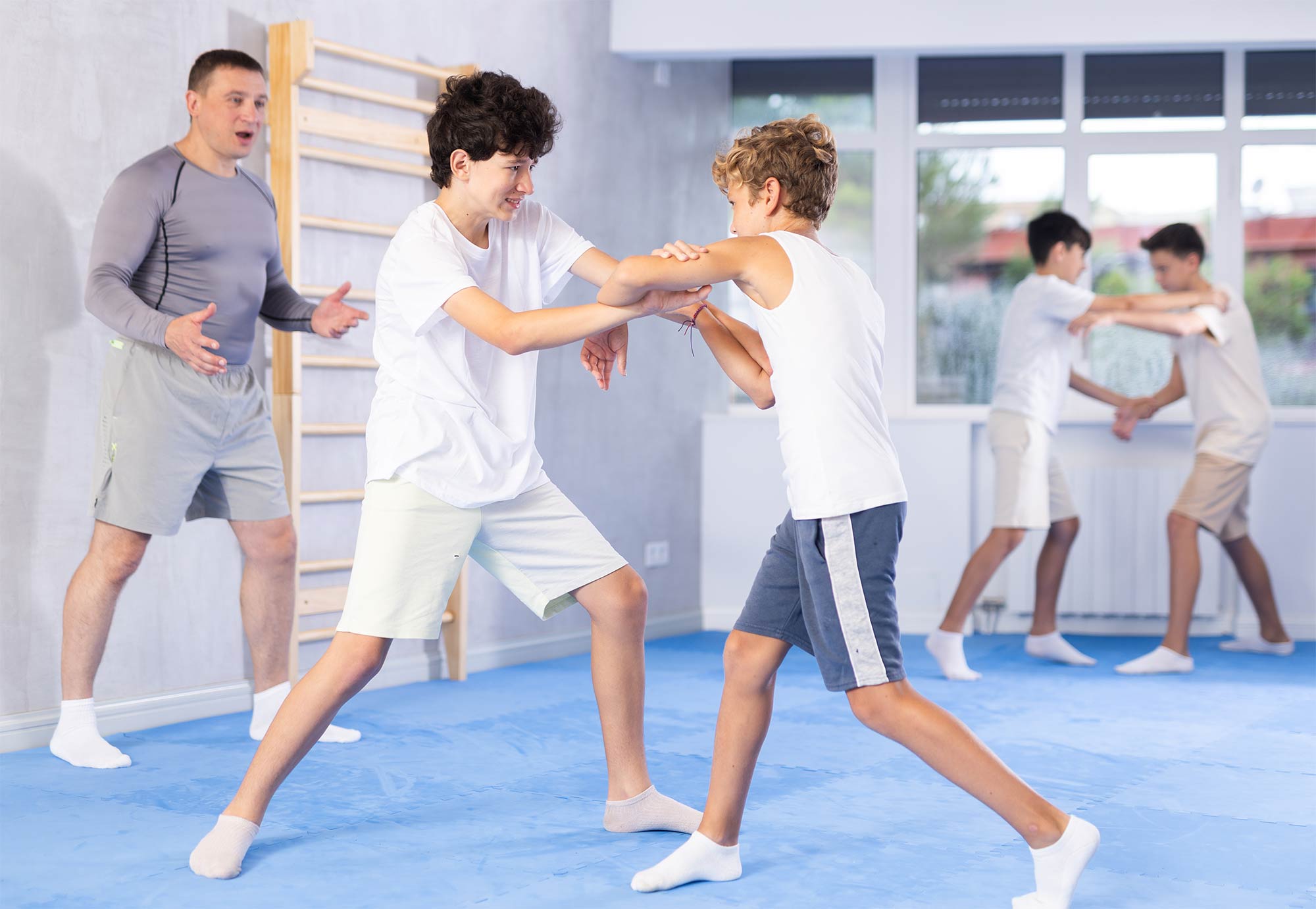 Zwei Jungs um alter von ca. 13 Jahren die konzentriert in lockerer Sportbekleidung Verteidigungs-griffe üben. Im Hintergrund steht ein männlicher Trainer im Alter von ca. 50 Jahren der ihnen Anweisungen gibt.